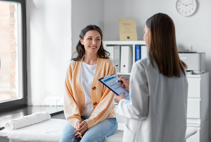 A woman sits on a medical examination table, smiling at a healthcare professional who holds a tablet, both within a modern clinic setting.