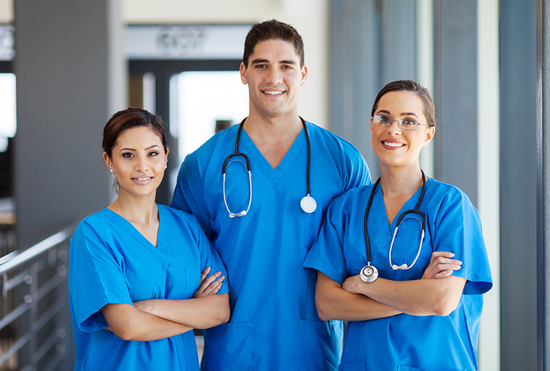 Three healthcare professionals posing confidently in front of an office building, with a woman standing between two men, all dressed in professional attire.