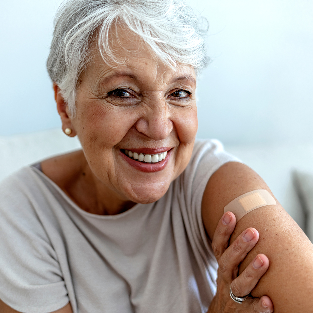 The image shows a woman with short hair smiling at the camera, wearing a white top, and has a visible bandage on her right arm, indicating she may have received a vaccination or medical treatment.